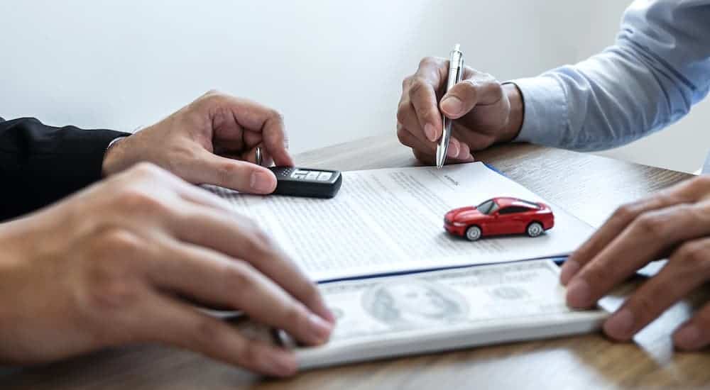 Two men are sitting at a desk with money, paperwork, car keys, and a red toy car.