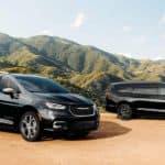 Two black 2021 Chrysler Pacificas are parked on dirt in front of tree-covered mountains.