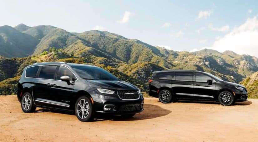 Two black 2021 Chrysler Pacificas are parked on dirt in front of tree-covered mountains.