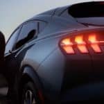 A silver 2021 Ford Mustang Mach-E is shown from the rear with the tail lights illuminated and the silhouette of a man standing next to it.