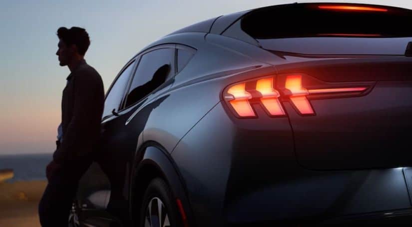 A silver 2021 Ford Mustang Mach-E is shown from the rear with the tail lights illuminated and the silhouette of a man standing next to it.