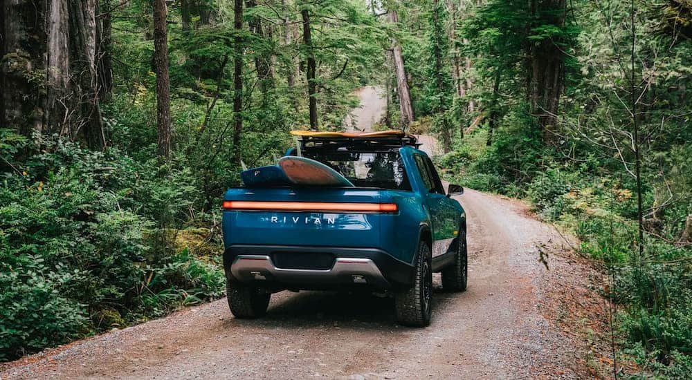 A blue 2021 Rivian R1T is driving through the woods with surf boards loaded on the roof and bed.