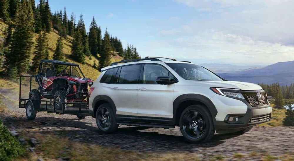 A white 2021 Honda Passport is towing a UTV with evergreen tress and hills in the background.