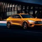 An orange 2021 Ford Mustang Mach-E is driving down a dark road with a modern building in the background.