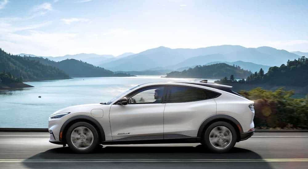 A popular Hybrid SUV, a white 2021 Ford Mustang Mach-E, is driving down the road with a lake and mountains in the background.