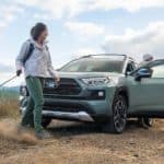 A pale green 2021 Toyota RAV4 Hybrid is parked on a hill with two women walking a dog.