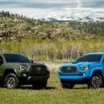 Two Toyota pickup trucks, a green and a blue 2021 Toyota Tacoma, are parked on grass in front of a hill.