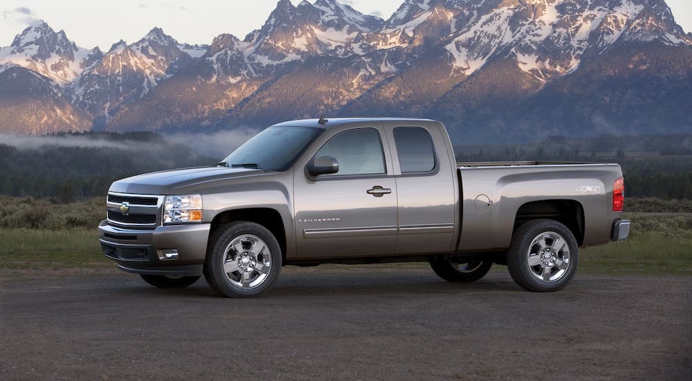 An older used Chevy diesel truck, a silver 2009 Chevy Silverado, is parked in front of snow-covered mountains.