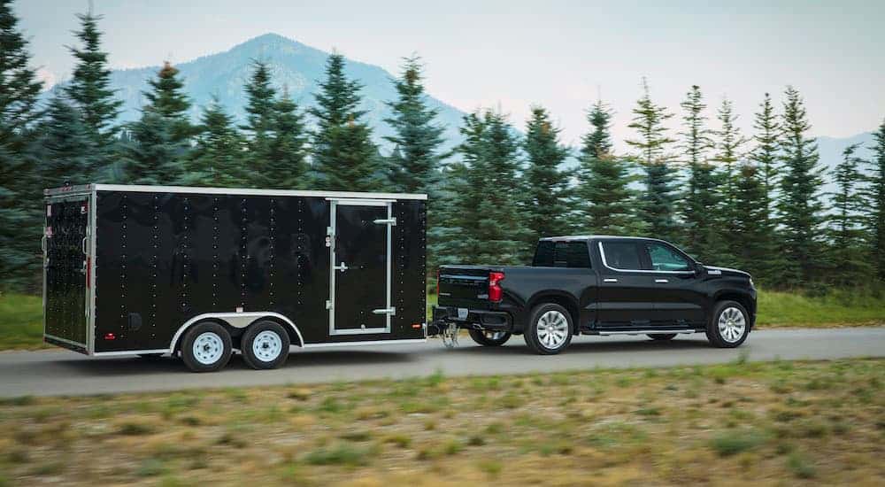 A black 2019 used Chevy Silverado is shown in profile towing a black enclosed trailer past a tree lined road.