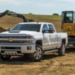 A used diesel truck, a white 2018 Chevy Silverado 2500HD with a trailer, is parked in front of construction equipment moving dirt piles.