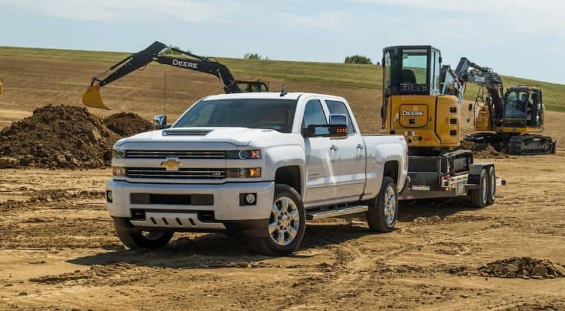 A used diesel truck, a white 2018 Chevy Silverado 2500HD with a trailer, is parked in front of construction equipment moving dirt piles.