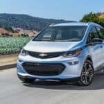 A silver 2021 Chevy Bolt EV is driving on a highway in front of mountains and fields.