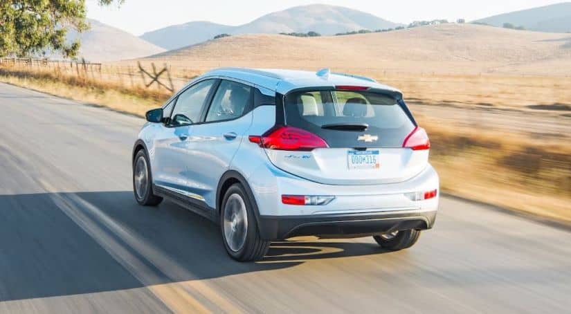 A silver 2021 Chevy Bolt EV is driving away on an empty highway towards mountains.