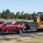 A red 2021 Chevy Silverado 3500 HD is towing construction equipment on a rural road.