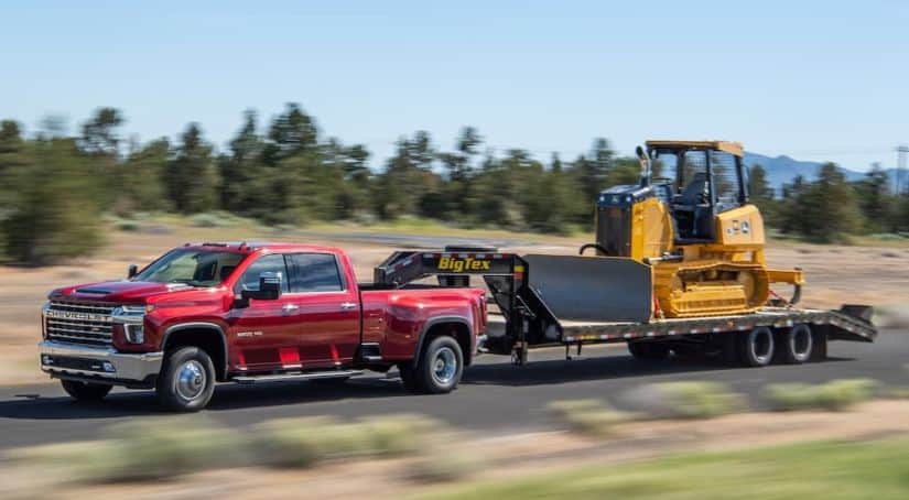 A red 2021 Chevy Silverado 3500 HD is towing construction equipment on a rural road.