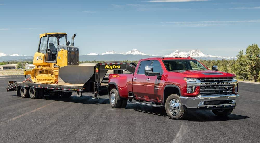 A red 2021 Chevy Silverado 3500 HD is in an open parking lot with heavy machinery on a trailer.