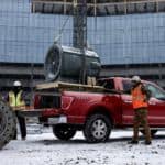 A large HVAC fan is being loaded into the bed of a red 2021 Ford F-150 at a construction site.