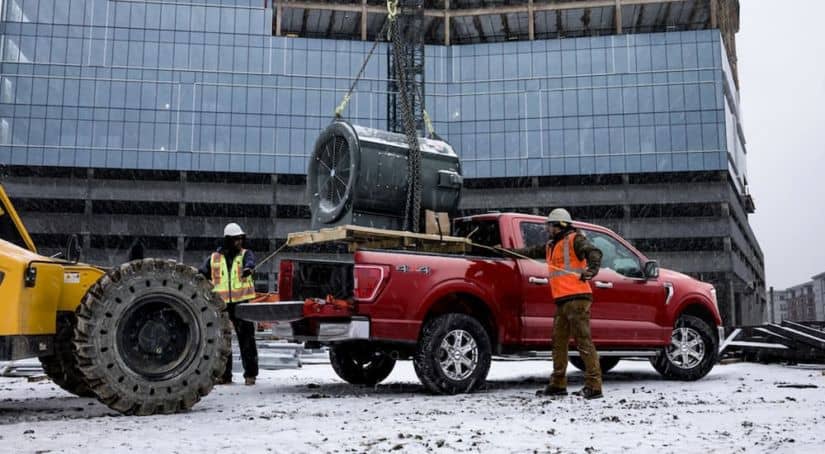 A large HVAC fan is being loaded into the bed of a red 2021 Ford F-150 at a construction site.