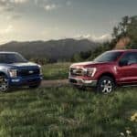 A blue and a red 2021 Ford F-150 are parked in a field in front of distant mountains.