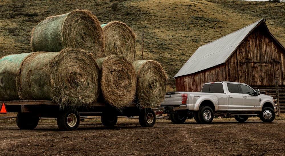 A silver 2021 Ford F-350 is towing a trailer with massive hay bales on a dirt driveway past a wood barn.