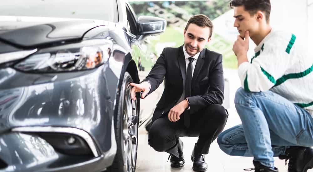 A salesman is showing a young man a tire on a grey car.