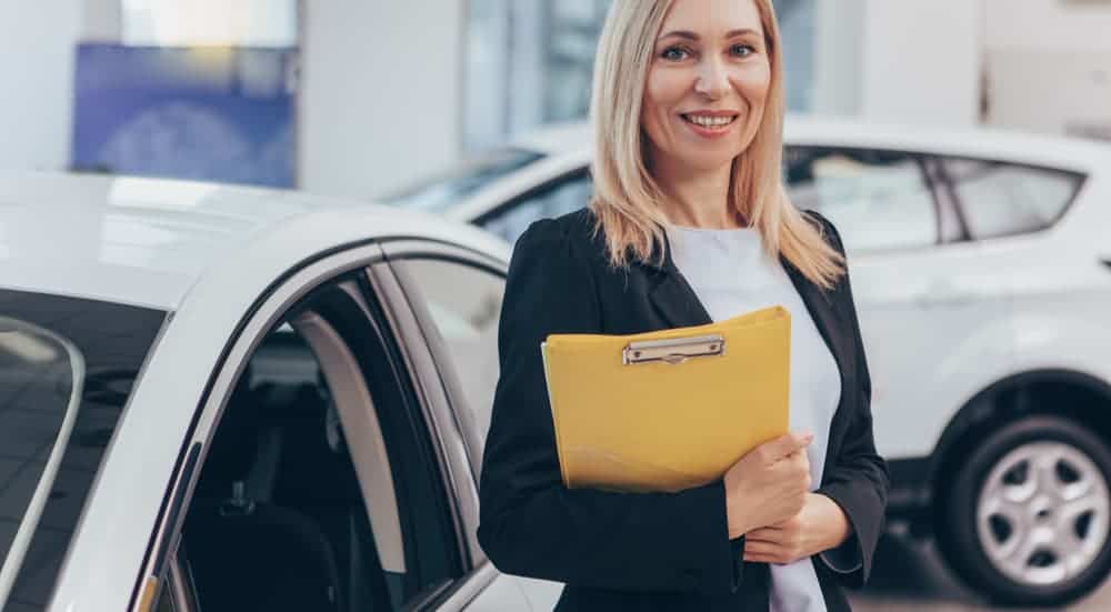 A saleswoman at a car dealership with a yellow clipboard is in front of two white cars.