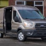 A brown 2021 Ford Transit cargo van is parked in front of a brick building with the side door open.