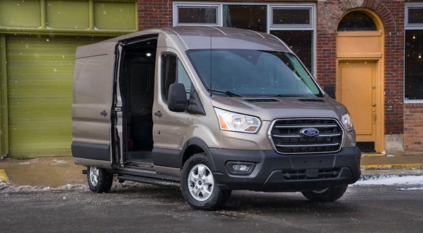 A brown 2021 Ford Transit cargo van is parked in front of a brick building with the side door open.