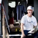 A worker in a hard hat is sitting in the open back door of a GMC Savana van with a laptop.