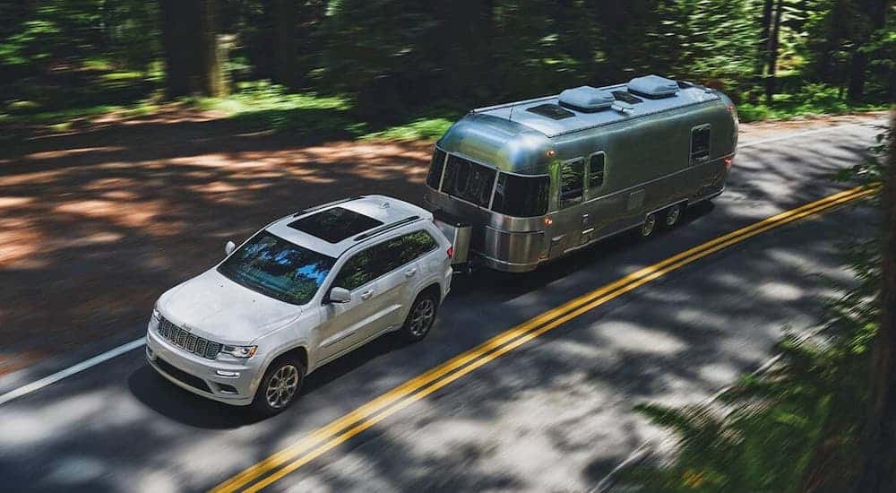 A high angle shows a white 2020 Jeep Grand Cherokee towing an Airstream camper on a woodland road.