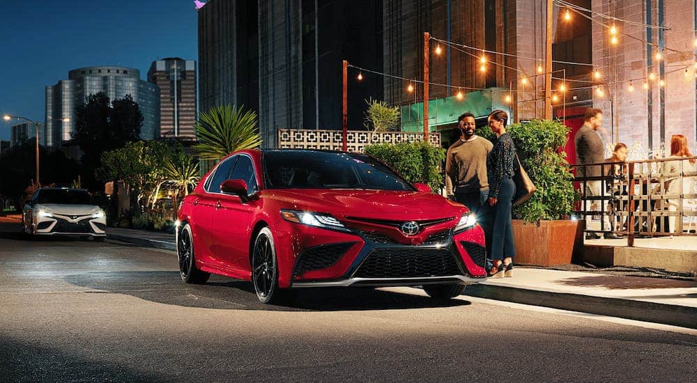 A couple is standing next to a red 2021 Toyota Camry Hybrid that is parked outside a restaurant at night.