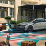 A silver 2021 Toyota Corolla Hybrid is parked on a colorful geometric mural while young adults drink coffee in the foreground.