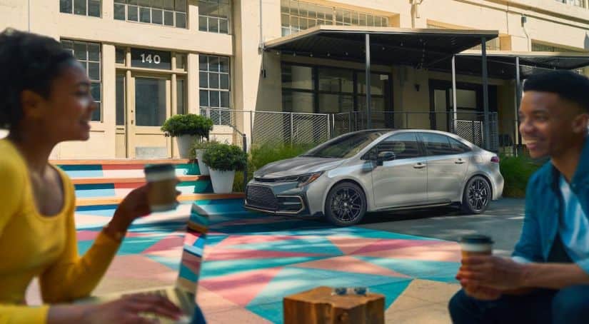 A silver 2021 Toyota Corolla Hybrid is parked on a colorful geometric mural while young adults drink coffee in the foreground.