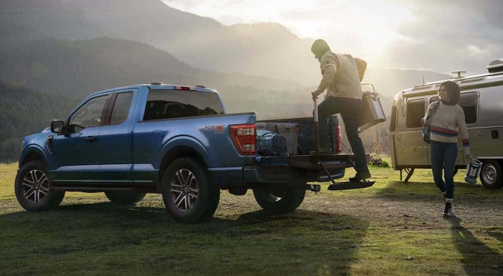 A person is using the tailgate step on a blue 2021 Ford F-150 in front of an Airstream and mountains.