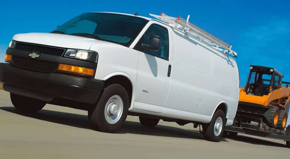 A white 2021 Chevy Express Cargo van is driving on a highway and towing construction equipment.