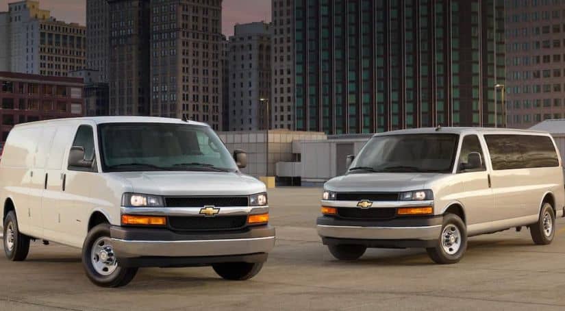 A white 2021 Chevy Express Cargo van and a silver Passenger van are parked on a parking garage in a city.