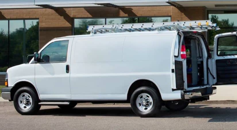A white 2021 Chevy Express Cargo Van with ladders on the roof is shown from the side with the rear doors open.