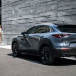A blue 2021 Mazda CX-30 is shown from the rear parked in front of a modern stone building with a couple approaching it.