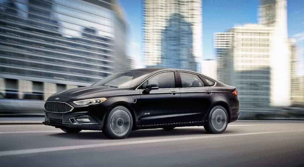 A black 2017 Ford Fusion Energi is speeding past a blurred city after leaving a used car dealer in Lexington.