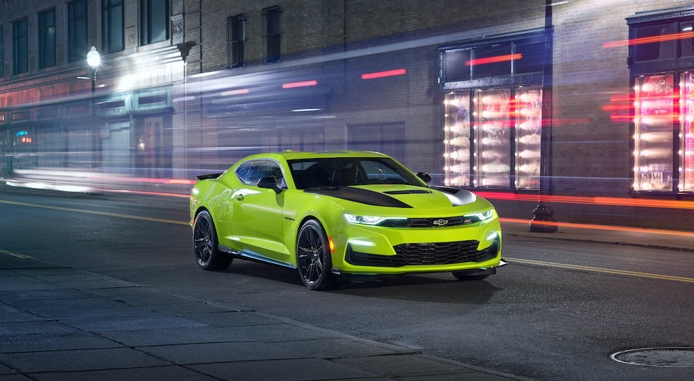 A green and black 2019 Chevy Camaro is driving past a blurred city building at night.