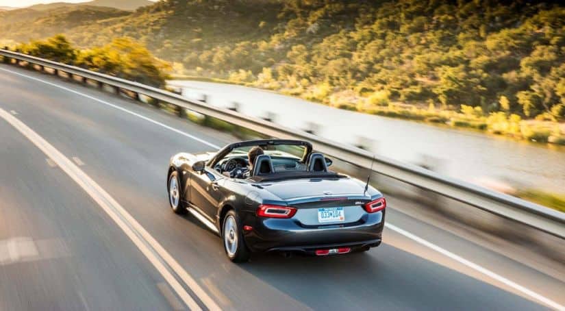 A gray 2017 Fiat 124 Spider convertible is shown from the rear driving on a highway past a river after leaving a used Fiat dealer.