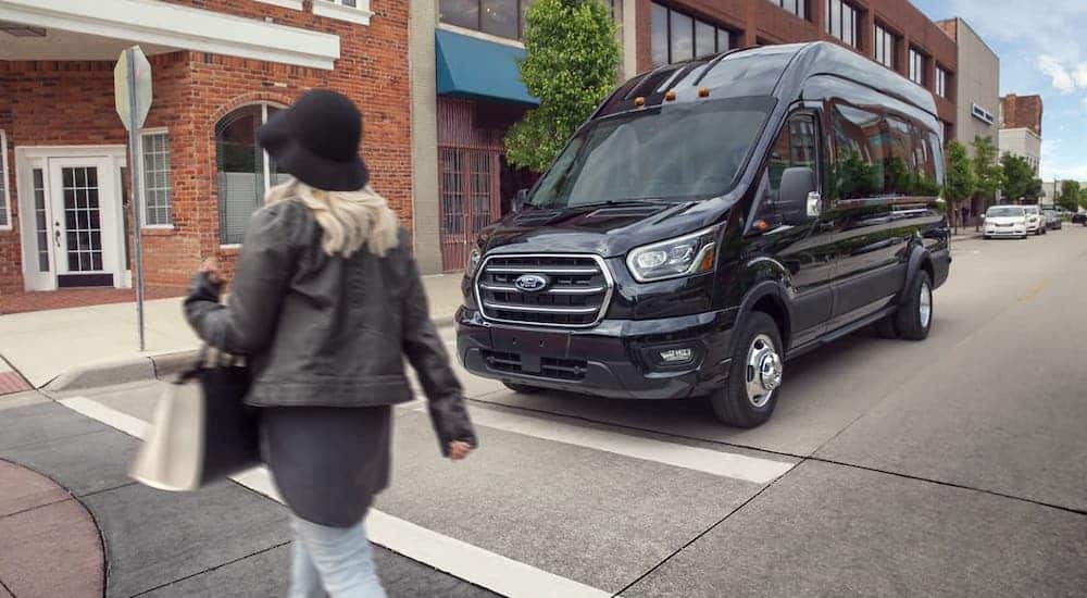 A woman is walking in a crosswalk in front of a parked black 2021 Ford Transit van, a popular used Ford commercial vehicle.