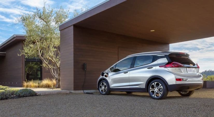 A silver 2021 Chevy Bolt EV is shown from behind while charging at a modern house.