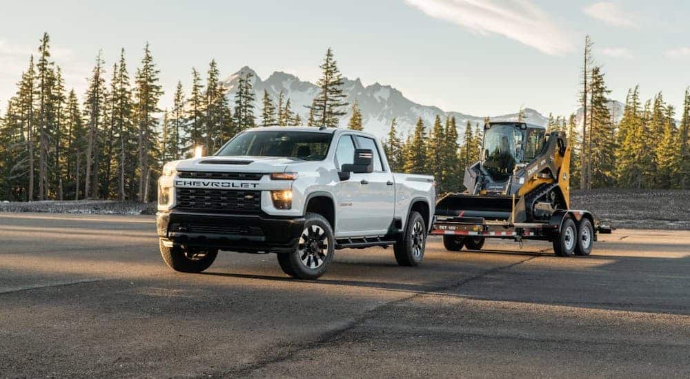 A white 2021 Chevy Silverado 2500 HD is towing construction equipment in front of distant trees and mountains.