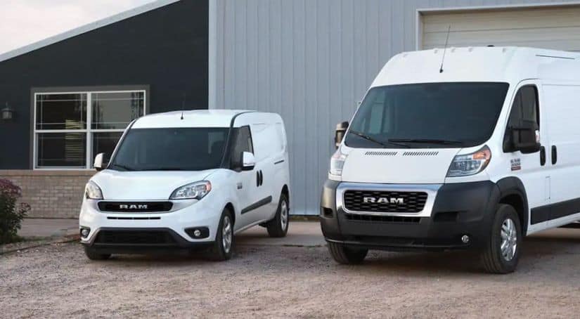 Two of the 2021 Ram commercial vehicles, both white 2021 Ram ProMaster and ProMaster City vans, are parked in front of a large metal building.