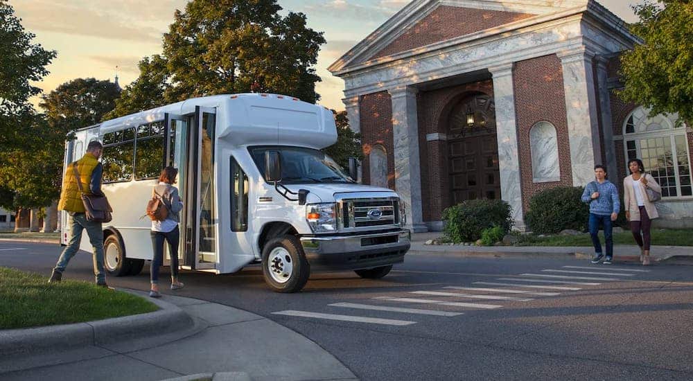 People are getting on a white 2022 Ford E-Series Cutaway shuttle that is parked in front of a red brick building.