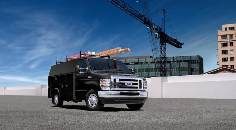 A black 2022 Ford E-Series Cutaway with storage containers is parked in front of a large metal crane.