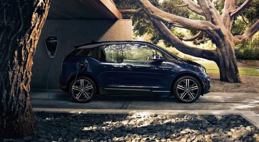 A dark blue 2021 BMW i3 is shown from the side charging under a brick structure in front of a large tree.