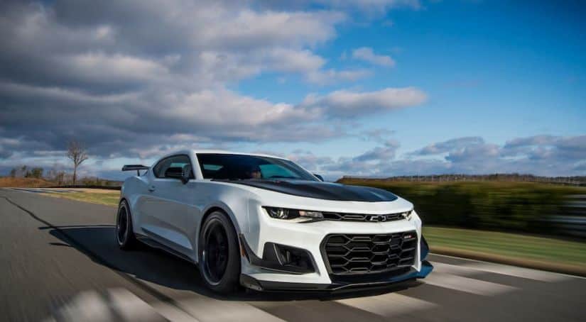 A white 2019 Chevy Camaro ZL1 is shown driving down the road with blue skies and clouds in the background.
