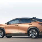 A bronze Nissan Ariya is shown from the side driving down the highway past a city.